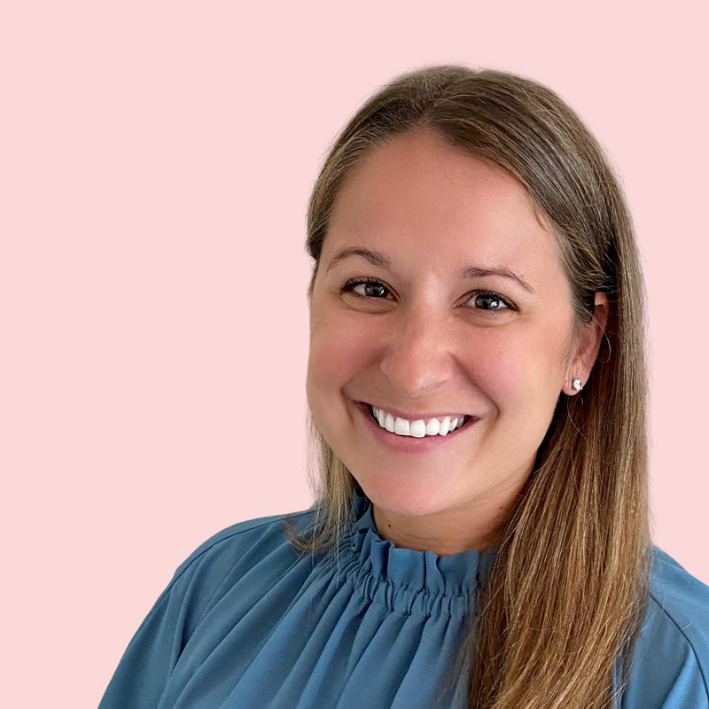 Woman smiling, wearing a blue blouse with a ruffled collar, against a plain pink background.