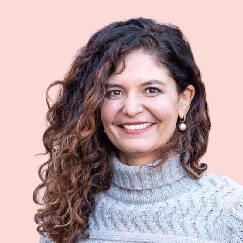 Woman with curly hair smiling, wearing a gray sweater against a pink background.