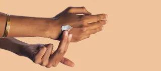 A person applies lotion to the back of their hand with a finger against a beige background.