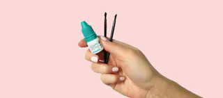 A hand holds a small bottle with a teal cap and two makeup brushes against a pink background.