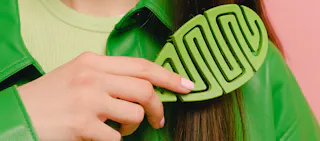A person with long brown hair holds a large green hair clip against a green jacket.