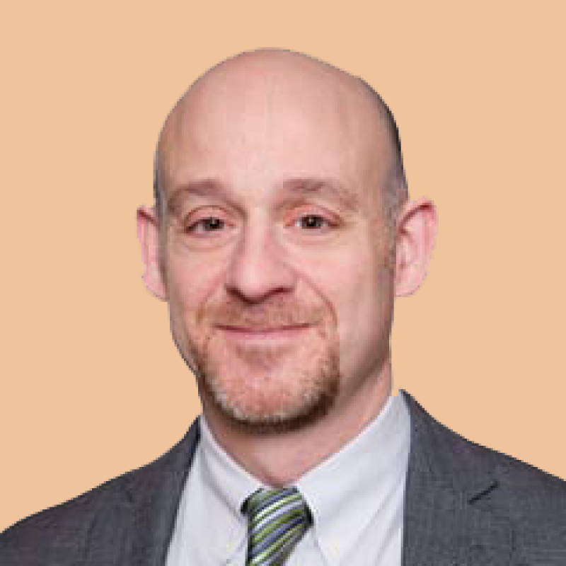 A bald man with a goatee wearing a suit and a striped tie, smiling at the camera, against a plain peach background.