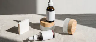 Skincare products with white labels and wooden lids on a beige cloth, featuring bottles with dropper caps and a rectangular soap bar.