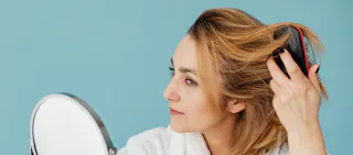 A woman brushes her hair while looking into a mirror against a blue background.