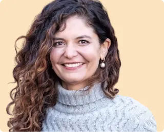 A person with long curly hair wearing a gray turtleneck sweater smiles against a beige background.