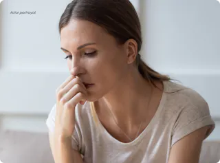 A woman in a light shirt sits thoughtfully with her hand against her chin, looking down.