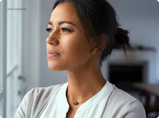 Woman with dark hair gazes out a window, wearing a white top and necklace.