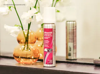 A bottle of Minoxidil topical aerosol foam for women on a bathroom counter, next to an orange vase with white flowers.