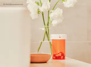 An orange pill bottle with red capsules sits on a counter near a small dish and a vase of white flowers.