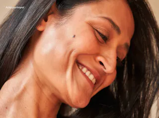 Close-up of a woman smiling with her head tilted down, showing a visible mole on her cheek. Her dark hair falls loosely around her face.