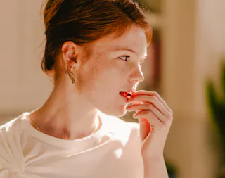 Person with red hair and freckles, wearing a white shirt and earrings, looking thoughtfully to the side while touching their lips.