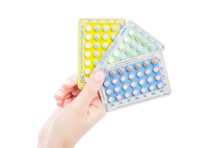 A hand holds three blister packs of contraceptive pills: yellow, green, and blue, each with differently colored tablets.