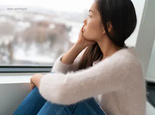 A woman sits by a window, gazing outside, with snow visible in the background. She rests her chin on her hand, wearing a light sweater and jeans.