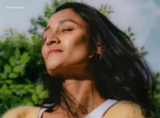 A person with long dark hair enjoys sunlight, eyes closed, with a blurred green leafy background.