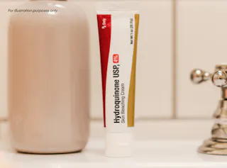 A tube of Hydroquinone USP, 4% Skin Bleaching Cream, is placed on a bathroom counter. A beige bottle and a chrome faucet are in the background. The text 