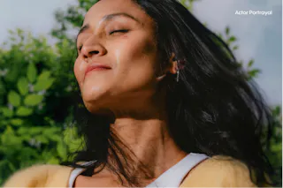 A person with long hair closes their eyes and tilts their head back, enjoying the sunlight. Trees and greenery are in the background.