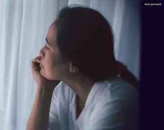 A person in a white shirt looks contemplatively out a window with sheer curtains.