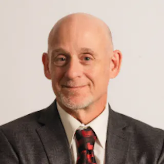 Bald man with a warm smile wearing a gray suit jacket, white shirt, and red plaid tie against a plain background.