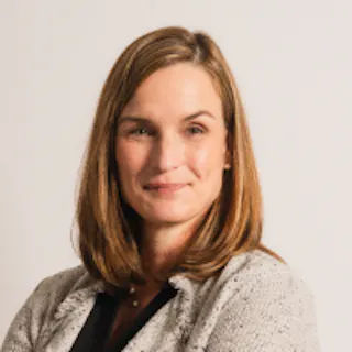 A person with shoulder-length brown hair and wearing a light gray blazer smiles at the camera against a plain background.