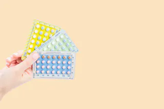 A hand holds three packs of birth control pills against a plain background.