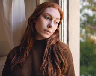 A woman with long red hair gazes out a window on a rainy day, wearing a brown sweater.