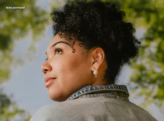 A person with curly hair and pearl earrings looks upward while standing outdoors, with green foliage in the background.