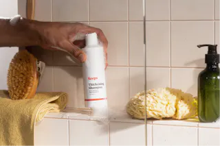 A hand reaches for a bottle of thickening shampoo on a shower shelf, surrounded by a bath brush, yellow towel, shower sponge, and soap dispenser.