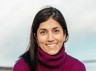 Woman with dark hair and a purple turtleneck smiling at the camera against a blurred outdoor background.