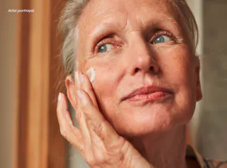 An older woman applies face cream, looking thoughtful.