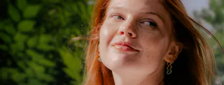 A person with red hair and earrings smiles softly, with a blurred green background.