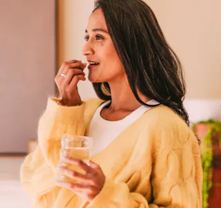 A woman in a yellow sweater holds a glass of water and a Loryna pill, preparing to take her medication.