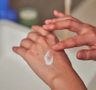 A person applies a small amount of hydroquinone cream to the back of their hand near a sink area.