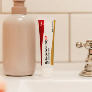 A tube of Hydroquinone Cream USP 2% sits on the bathroom sink beside a soap dispenser, framed by a tiled wall in the background.