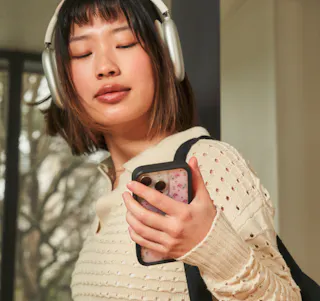 A person wearing headphones and a beige sweater holds a smartphone with a floral case, glancing at an article on Tretinoin Cream.
