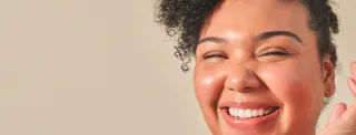 A person with curly hair, radiant from their Tretinoin Cream routine, smiles broadly, showing their teeth against a light background.