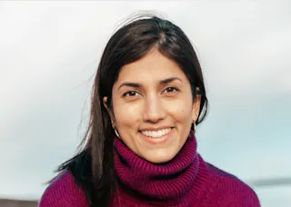 A person with long dark hair smiles at the camera, wearing a purple turtleneck sweater. The background is a blurred sky.