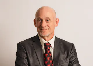 Bald man in a gray suit and red patterned tie, smiling slightly, against a plain white background.