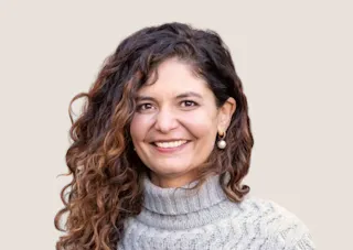 Woman with curly hair wearing a gray turtleneck sweater, smiling against a plain background.