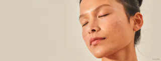 A person with closed eyes and freckles, head tilted slightly back, hair tied up, against a neutral background, enjoys the refreshing effects of Tretinoin Cream.