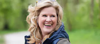 Person with wavy blonde hair, wearing a blue vest, smiling outdoors with greenery in the background.