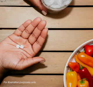 A person holds two tablets in their hand, next to a glass with ice and a bowl of assorted small peppers on a wooden surface.