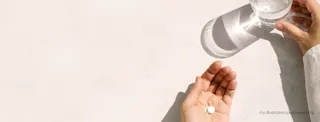 A person holds two pills in one hand and a glass of water in the other on a white surface.