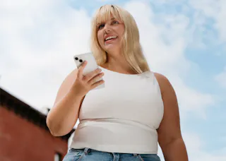 A person with long blond hair holds a smartphone outdoors, smiling and looking up. They're wearing a white sleeveless top and jeans against a background of a blue sky and a brick building.