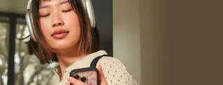 Woman with headphones looks at her smartphone; wears a beige textured sweater and has shoulder-length hair.
