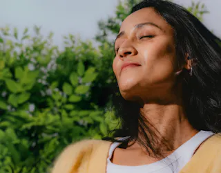 A person stands outdoors with eyes closed, enjoying the sunlight. Trees and green foliage are visible in the background.