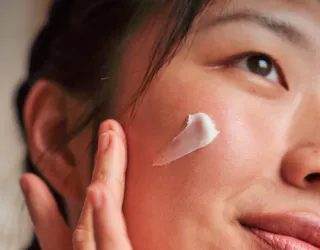 A person applies a dab of white cream to their cheek with their fingers.