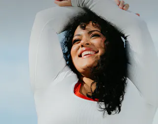 A person with curly hair and a bright smile stretches their arms over their head, wearing a white top against a clear sky background.