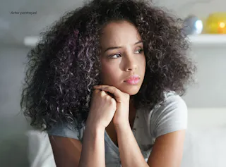 A woman with curly hair sits indoors, resting her chin on her hands and gazing pensively to the side.