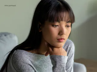A young woman with long dark hair sits indoors, resting her chin on her hand and looking down with a thoughtful, serious expression.