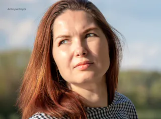 A woman with long auburn hair wearing a black and white checkered top looks slightly upward outdoors on a sunny day.
